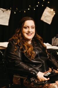 Carly Neis smiles at the camera, sitting in her wheelchair, she's wearing a black leather jacket and gold sparkle skirt. Behind her are string lights with sheet music pages hanging from the lights. There is also a desk with more sheet music.
