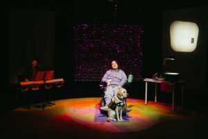 Carly Neis sits in her wheelchair center stage with her support dog Gilmore sitting at her feet. The light wall behind her has a soft red twinkling as Carly and Gilmore and lit up with a spotlight showing the multi-colour rug on the stage.