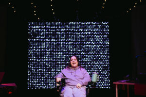 Carly is center stage wearing a purple sweat suit and sitting in her wheelchair. Behind her is a twinkling wall of lights, Carly is looking out past the camera to the audience.