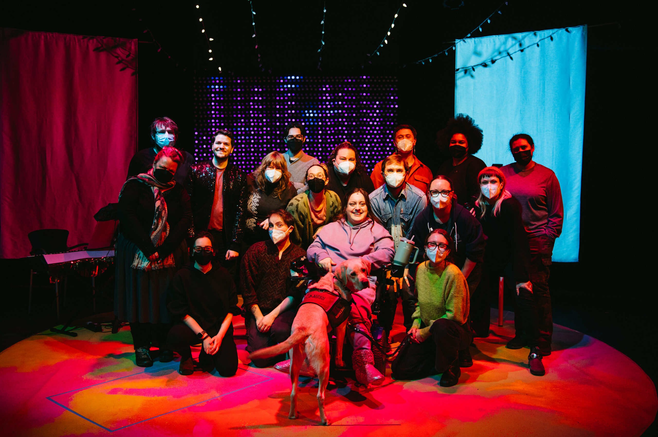 In My Own Little Corner cast and crew all center stage. Carley Neis is center with support dog Gilmore. She is surrounded by 16 team members wearing face masks. There are string lights overhead and a light wall behind the group.