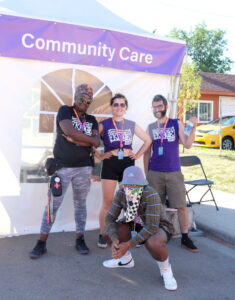 The Community Care team stands outside their tent that is labelled Community Care with a purple banner. Manager, Shima, stands to the left wearing a black shirt, and the other team members wear a purple shirt.