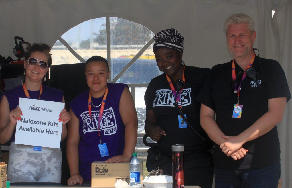 Community Care Team stands together smiling inside their tent. Two staff members stand to the right in black shirts, and two volunteers stand to the left in purple shirts. One of the volunteers is holding a sign that says: naloxone kits available here.