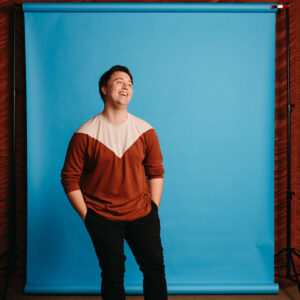 Mac Brock stands in front of a blue backdrop wearing a brown and white sweater.