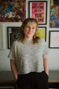 Jessica smiles at the camera while standing in front of a wall of framed art. She wears a black and white striped top with black pants, her hands are in her pockets. She has long earrings and an auburn and blonde fringe haircut.