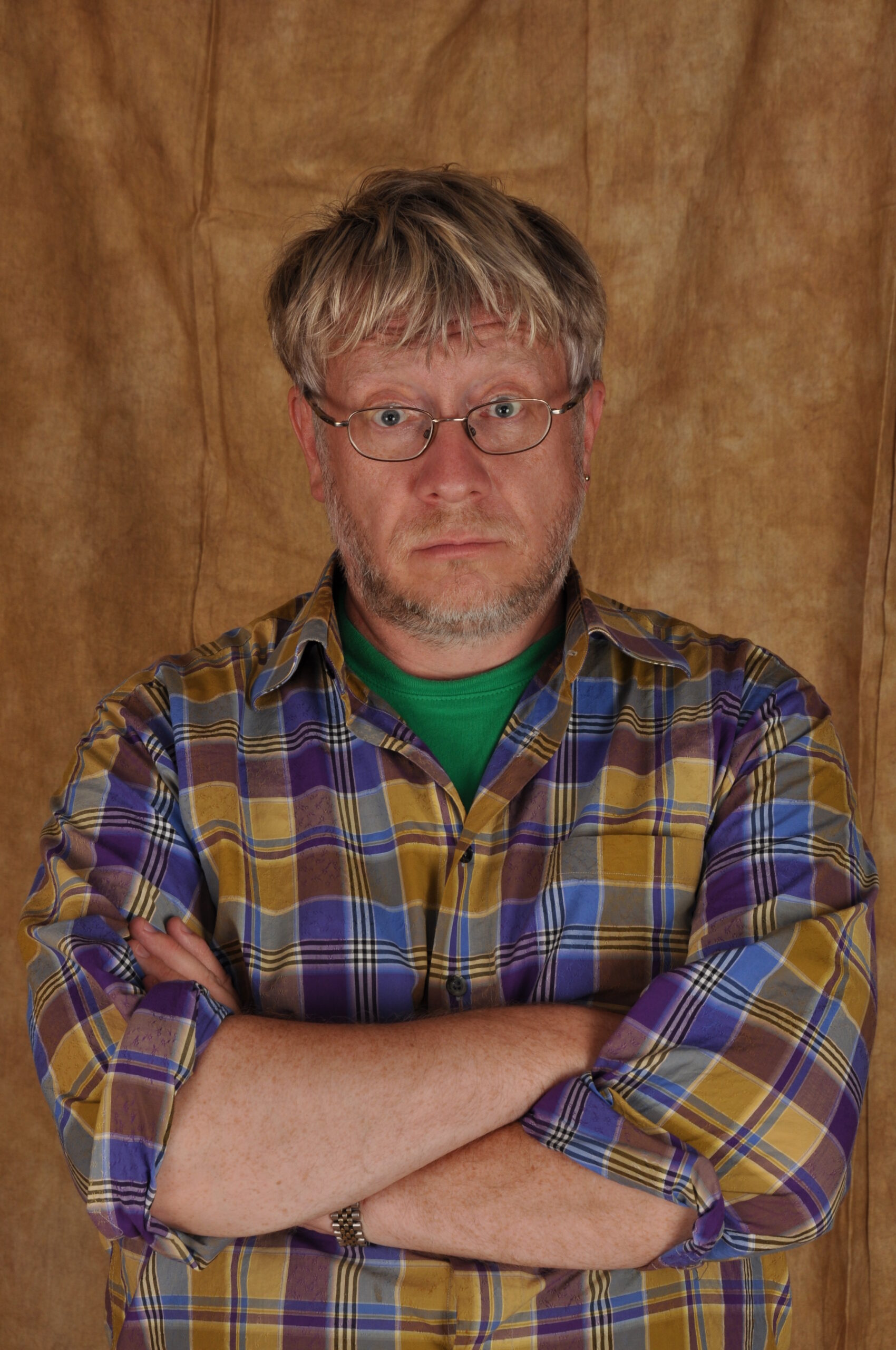 Older man with glasses and a plaid shirt crosses his arms in front of a brown backdrop.