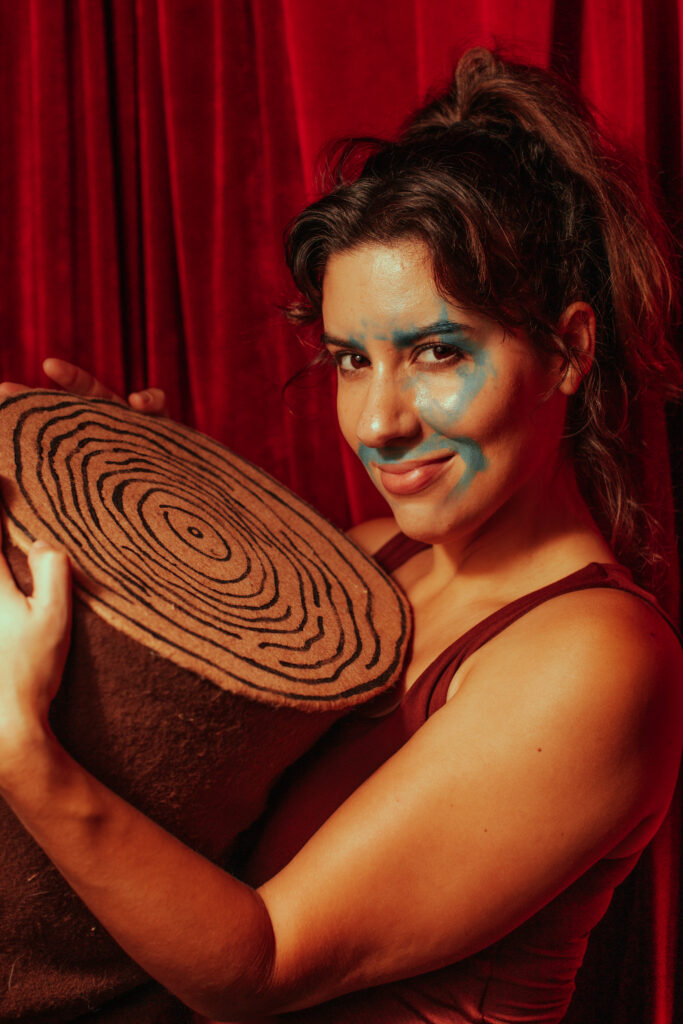 Artist Natasha Mercado is holding a prop tree stump while looking at the camera and smiling.
