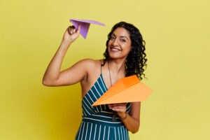 Artist Aliya Kanani is standing up with a big smile, holding two paper airplanes.
