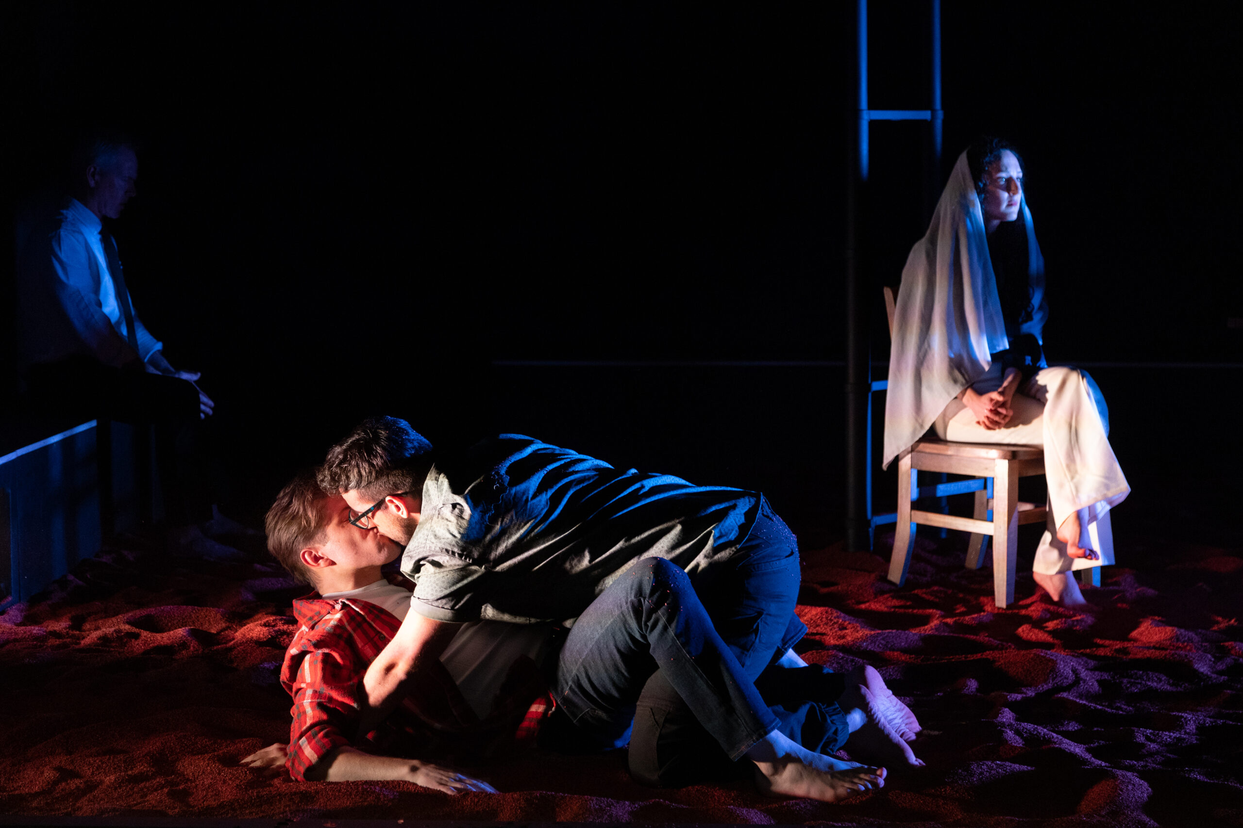 Production photo of Hooves featuring two men on top of each other, kissing. In the background, a woman sits on a chair in dramatic lighting.