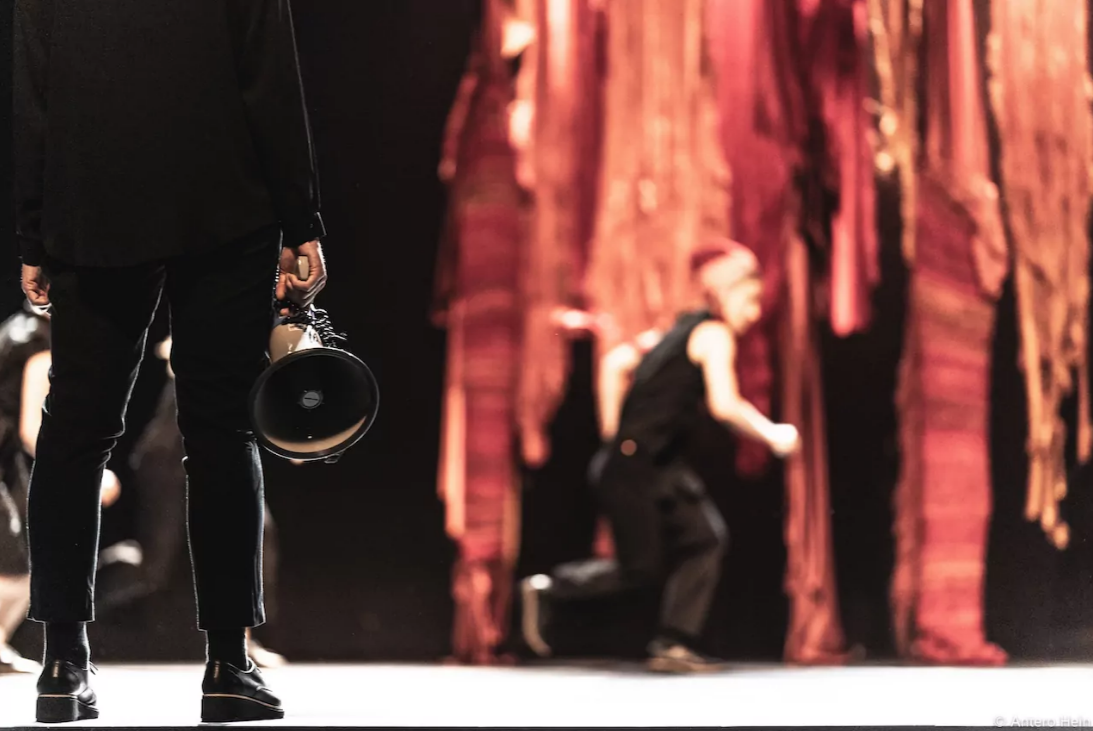 Photo from the production featuring someone standing in the foreground with a megaphone. In the background - a dancer runs between hanging fabric.