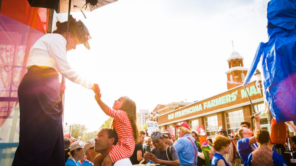 The festival in full-swing outside the Old Strathcona Farmers Market. A woman in Klondike wear stands tall above the crowd, carrying a sun umbrella and reaching out towards a small child in red-and-white stripes who is perched on a man's shoulders.