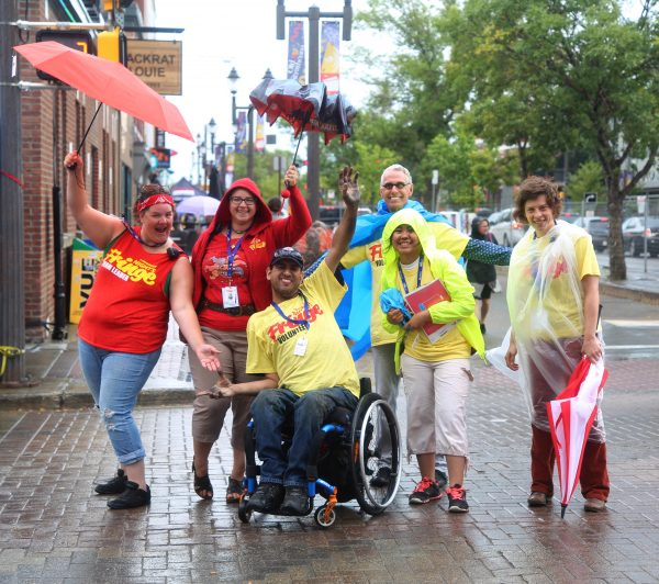 Six Fringers in the rain; they are wearing plastic ponchos, carrying umbrellas. One is front-and-centre in a wheelchair; they are on a brick road in Old Strathcona.