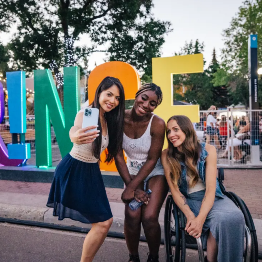 Three friends taking a selfie with a large Fringe word sculpture in the background.