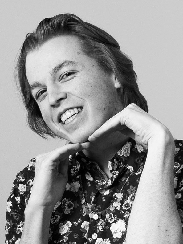 Bradley King in black-and-white smiles at the camera, wearing a flower-print shirt, framing his grinning face with the tops of his hands.