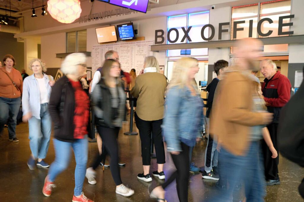 A blurry group of people walking through a busy lobby. A large sign that says "Box Office" is in the background.