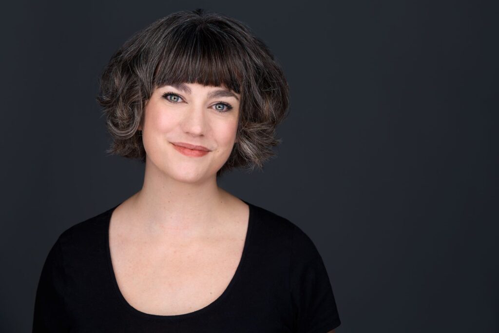 Megan Dart in a black shirt smiles at the camera, standing in front of a dark grey backdrop.