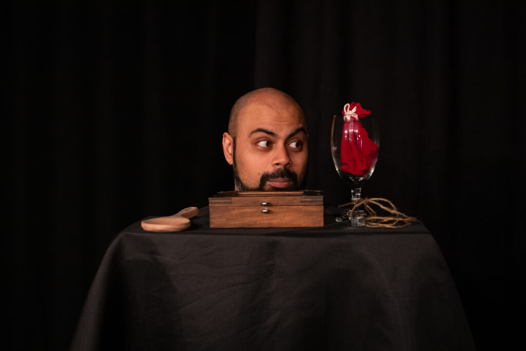 Shawn pokes his head above a table. There is a wooden box and a glass cup with a red napkin inside it.