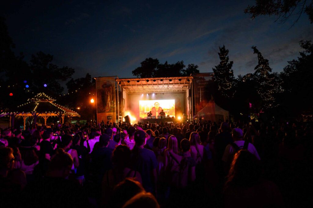 A night-time crowd bathed in magenta light, turned towards a DJ stage that glows orange, trees and a gazebo clad in twinkling lights framing the evening.